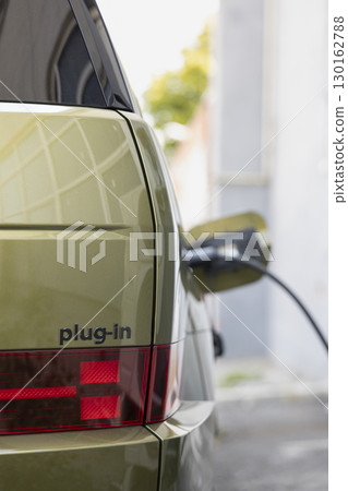 Close up of red tail light and plug in badge on electric vehicle charging at public station. Electric car plugged into charger, reducing greenhouse gas emissions, sustainable transport concept Close up of red tail light and plug in badge on electric vehicle charging at public station. Electric car plugged into charger, reducing greenhouse gas emissions, sustainable transport concept 130162788