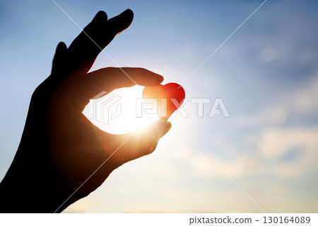 A silhouette of a woman holding a red heart, a symbol of precious life and love, against a blue sky and bright sunlight background. A silhouette of a woman holding a red heart, a symbol of precious life and love, against a blue sky and bright sunlight background. 130164089