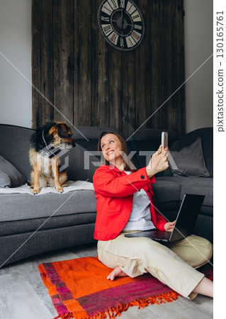 Woman working remotely with laptop, taking a selfie with her dog at home. Happy freelancer enjoying break with pet, sitting on floor in cozy living space, casual digital lifestyle 130165761