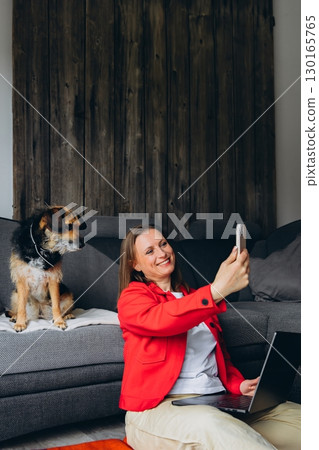 Woman working remotely with laptop, taking a selfie with her dog at home. Happy freelancer enjoying break with pet, sitting on floor in cozy living space, casual digital lifestyle Woman working remotely with laptop, taking a selfie with her dog at home. Happy freelancer enjoying break with pet, sitting on floor in cozy living space, casual digital lifestyle 130165765