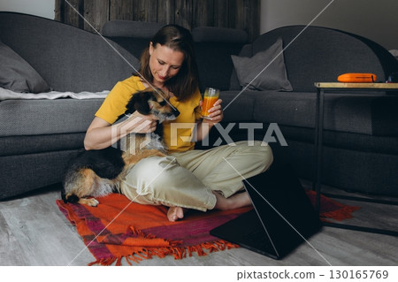 Smiling woman working remotely with laptop, holding juice, sitting on floor with her dog. Concept of freelance work, healthy lifestyle, and connection with a pet at home Smiling woman working remotely with laptop, holding juice, sitting on floor with her dog. Concept of freelance work, healthy lifestyle, and connection with a pet at home 130165769