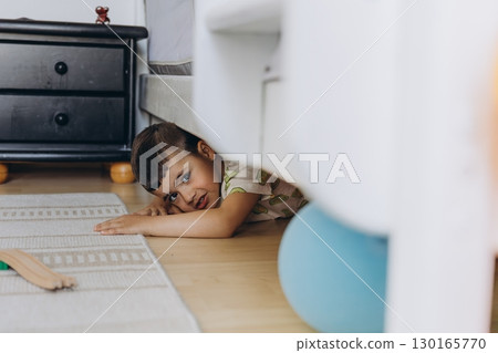 Close-up of curious young boy peeking out from under his bed in a cozy children room. Playful and imaginative moment at home. Natural light, warm tones, candid childhood scene 130165770