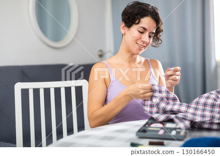 Positive young adult woman stitching on button to shirt 130166450