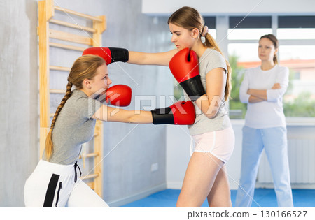 Two teenage girls in sportswear are engaged in boxing sparring in the gym under guidance of trainer in gym 130166527