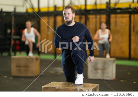 Young bearded man performing step-ups on box during group workout 130166603