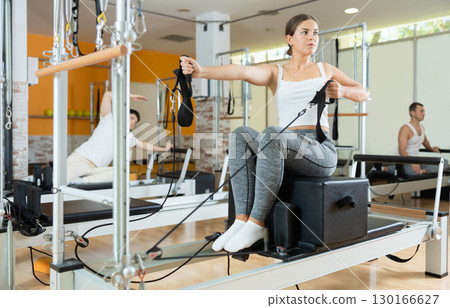 Girl in gym with help of rope reformer performs exercises to strengthen muscles of her arms. 130166627
