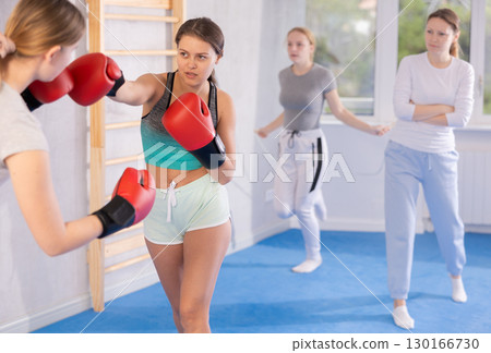 Two teenage girls in sportswear are engaged in boxing sparring in the gym under guidance of trainer in gym 130166730