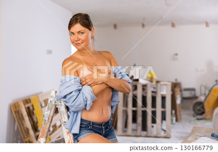 Seminude woman in unbuttoned shirt covering breasts with hands in room during renovation 130166779