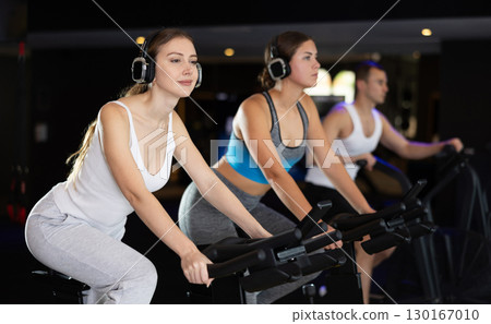 Girl in earphone performs exercises with stationary bicycle in gym Girl in earphone performs exercises with stationary bicycle in gym 130167010