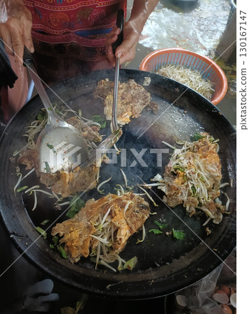 Person cooking large pieces of fried chicken with bean sprouts and herbs on hot griddle, using tongs and spatula, in outdoor setting 130167147
