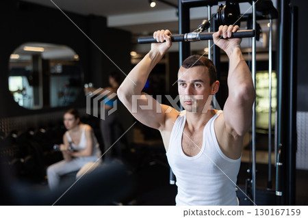 Slender athletic man work out muscles of arms and shoulders on simulator in gym of fitness club Slender athletic man work out muscles of arms and shoulders on simulator in gym of fitness club 130167159