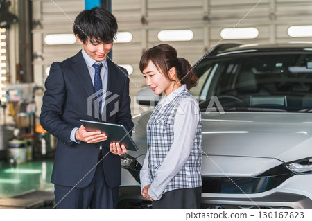 A male salesman and a female office worker looking at a tablet in front of a car in a repair shop pit 130167823
