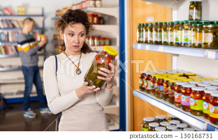 Female shopper selects and buys a jar of pickled cucumbers and tomatoes in grocery supermarket 130168731