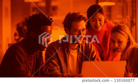 Diverse group of young professionals collaborating happily around laptop in warm office environment, focused on teamwork and creativity 130168877