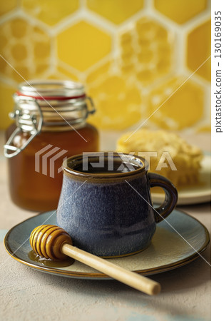 Coffee in dark ceramic cup with honey and jar of honey in background, honey dipper on plate, honeycomb, and honey spoon, set against honeycomb patterned yellow wall Coffee in dark ceramic cup with honey and jar of honey in background, honey dipper on plate, honeycomb, and honey spoon, set against honeycomb patterned yellow wall 130169035