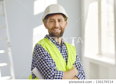 Portrait of positive, handsome young male construction worker in hard hat, smiling at the camera while working on construction site. 130170514