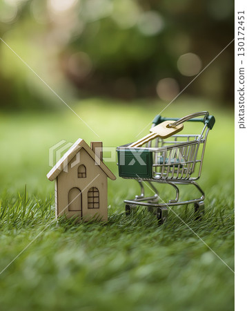 Small wooden house model next to miniature shopping cart filled with small shovel and gardening tools, set on green grass with blurred natural background, symbolizing home gardening or real estate 130172451
