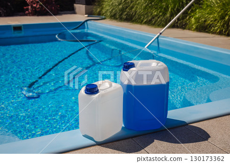 Two chemical canisters for pool treatment placed on the poolside edge, with a vacuum hose and clean blue water in the background on a sunny day. 130173362