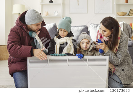 Family in warm clothes sitting by electric heater, trying to stay warm in cold room during winter. Family in warm clothes sitting by electric heater, trying to stay warm in cold room during winter. 130173633