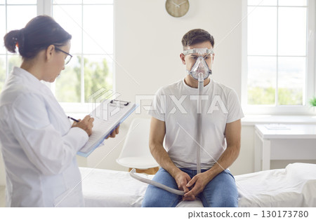 Male patient sitting on hospital bed in oxygen mask while doctor with clipboard recording some data. 130173780