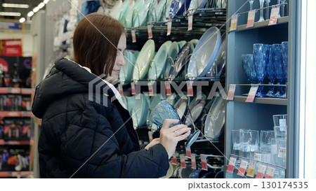 Young woman shopping for kitchenware, examining a blue glass in a supermarket, surrounded by various plates and glasses on display 130174535