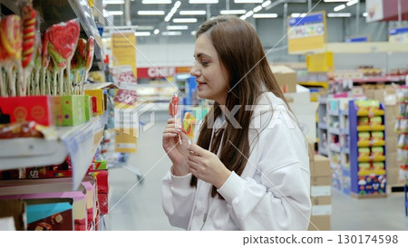 Woman deciding between two lollipops in a brightly lit candy aisle of a supermarket, surrounded by colorful sweets and promotions 130174598