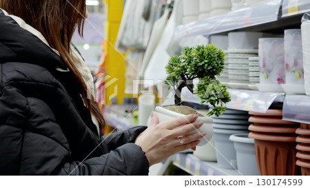 Customer shopping for houseplants in supermarket garden center, holding small bonsai tree and browsing selection of pots and home decor 130174599