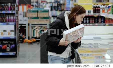 Young woman carefully examining a frozen pizza box in the supermarket freezer section, making grocery shopping decisions Young woman carefully examining a frozen pizza box in the supermarket freezer section, making grocery shopping decisions 130174601