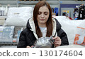 Young woman carefully selecting a stainless steel saucepan while shopping for kitchenware in a supermarket, surrounded by various household items 130174604