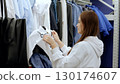 Young woman shopping for clothes, examining a white shirt in a fashion retail store, surrounded by various garments on hangers 130174607
