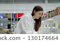 Young woman carefully selecting groceries from refrigerated shelves, navigating the frozen food aisle of a brightly lit grocery store 130174664