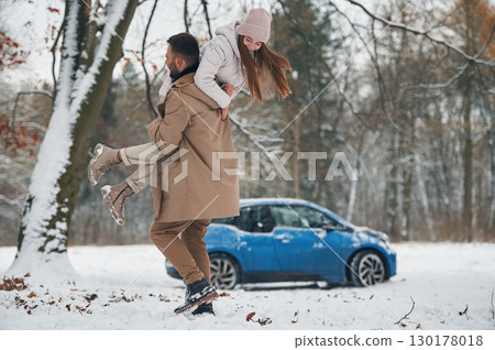 Playful mood. Happy couple having a walk in winter forest. Blue car is parked 130178018