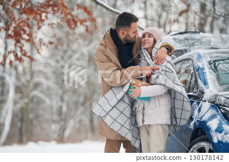 Man is holding the towel to warm the woman. Happy couple having a walk in winter forest. Blue car is parked 130178142