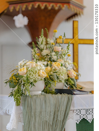 Beautiful floral arrangement with roses and hydrangeas decorates a church altar, complemented by a fabric drape. The setup includes a cross and open book, symbolizing a spiritual setting 130178310