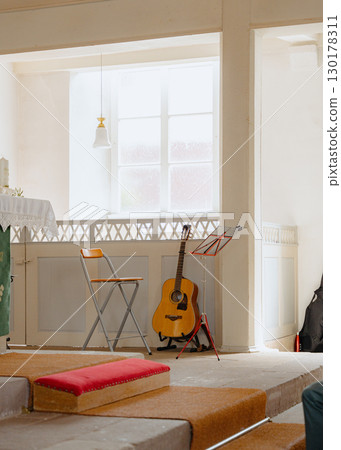Sunlit room with an acoustic guitar resting on a stand beside a folding chair and sheet music stand. The setting suggests a peaceful atmosphere, ideal for music practice or performance 130178311