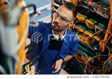 Looking up, management of cables. Young man is working with internet equipment and wires in server room 130178336