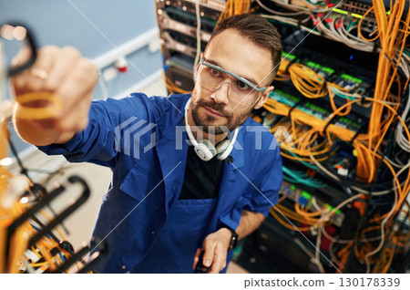 Looking up, management of cables. Young man is working with internet equipment and wires in server room Looking up, management of cables. Young man is working with internet equipment and wires in server room 130178339