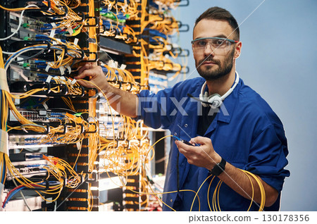 Looking to the camera. Young man is working with internet equipment and wires in server room Looking to the camera. Young man is working with internet equipment and wires in server room 130178356