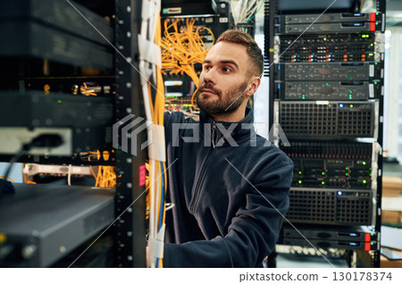 Black clothes. Young man is working with internet equipment and wires in server room Black clothes. Young man is working with internet equipment and wires in server room 130178374