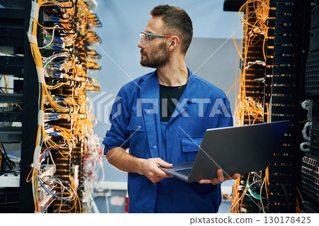 Holding laptop. Young man is working with internet equipment and wires in server room Holding laptop. Young man is working with internet equipment and wires in server room 130178425