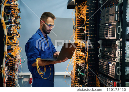 With laptop in hands. Young man is working with internet equipment and wires in server room 130178430