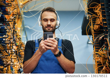 Headphones and smartphone. Young man is working with internet equipment and wires in server room 130178511