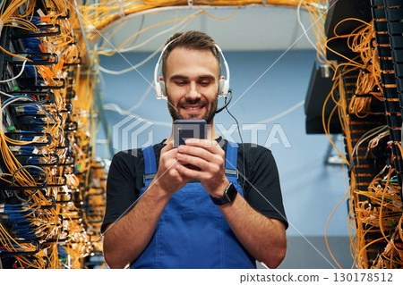 Headphones and smartphone. Young man is working with internet equipment and wires in server room 130178512