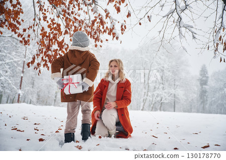 Girl is holding gift box for woman, behind her. Mother and her daughter is on the winter meadow and forest 130178770
