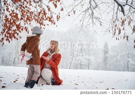 Girl is holding gift box for woman, behind her. Mother and her daughter is on the winter meadow and forest 130178771