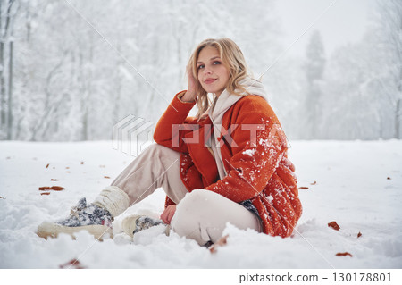 Beautiful woman in red warm jacket is sitting on the winter field 130178801