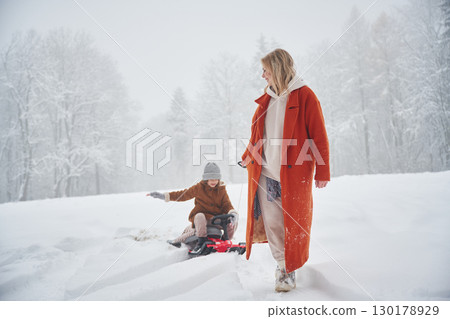 A woman pulls a sled. Mother and her daughter is on the winter meadow and forest 130178929