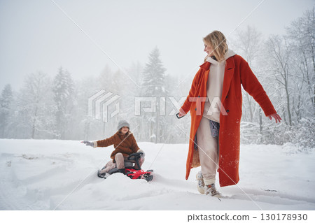 A woman pulls a sled. Mother and her daughter is on the winter meadow and forest A woman pulls a sled. Mother and her daughter is on the winter meadow and forest 130178930