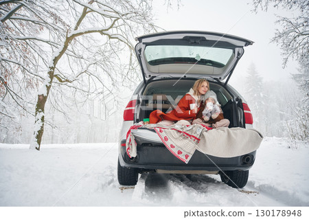 Sitting, with warm drink. Woman with her daughter is on the trunk of a car in the winter meadow and forest 130178948
