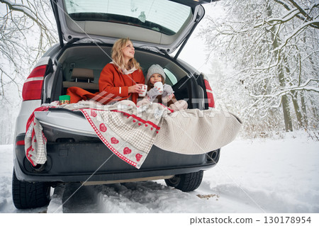 Drinking from the cup. Woman with her daughter is on the trunk of a car in the winter meadow and forest 130178954